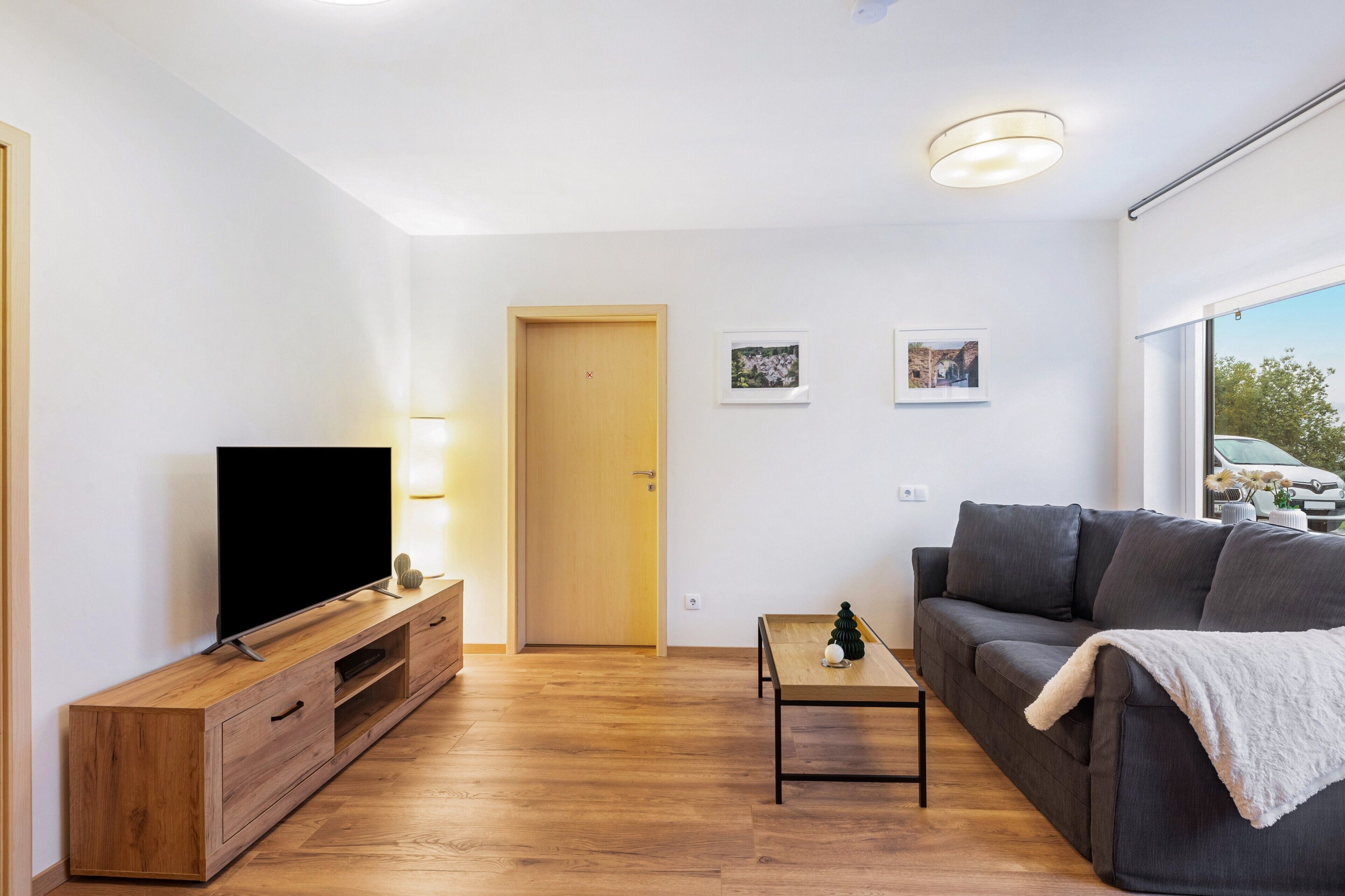 A modern living room with a flat-screen TV, wooden entertainment unit, grey sofa, and a small table with a lamp.