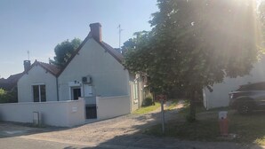 Un bâtiment blanc avec un toit en pente, une cheminée et une petite porte. On y voit une enseigne avec du texte, un arbre et une voiture garée.