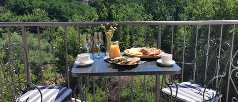 A balcony with a table set for breakfast, including bread, fruit, and drinks, overlooking a forested landscape.
