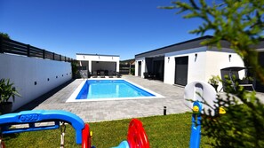 A modern outdoor pool area with a clear blue pool, a covered seating area, and a white building with a garage.