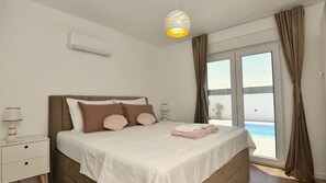 A bedroom with a bed, bedside tables, a lamp, a ceiling light, and a view of a swimming pool through large windows.