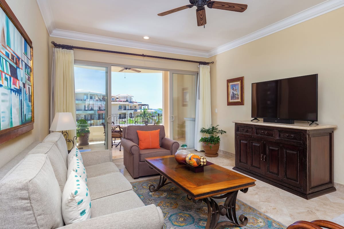 A living room with a sofa, a coffee table, a television on a cabinet, and a view of the outside.