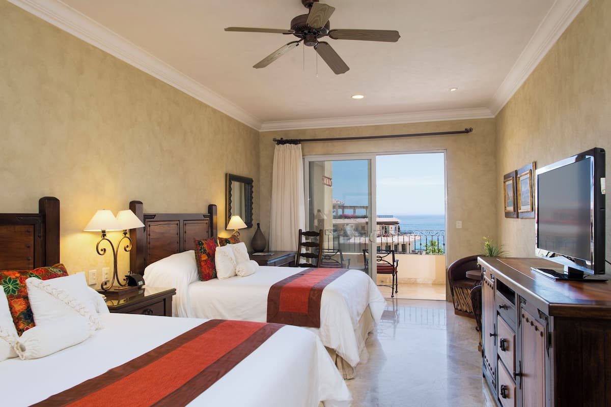 A hotel room with two beds, a TV, a ceiling fan, and a balcony with a view of the ocean.