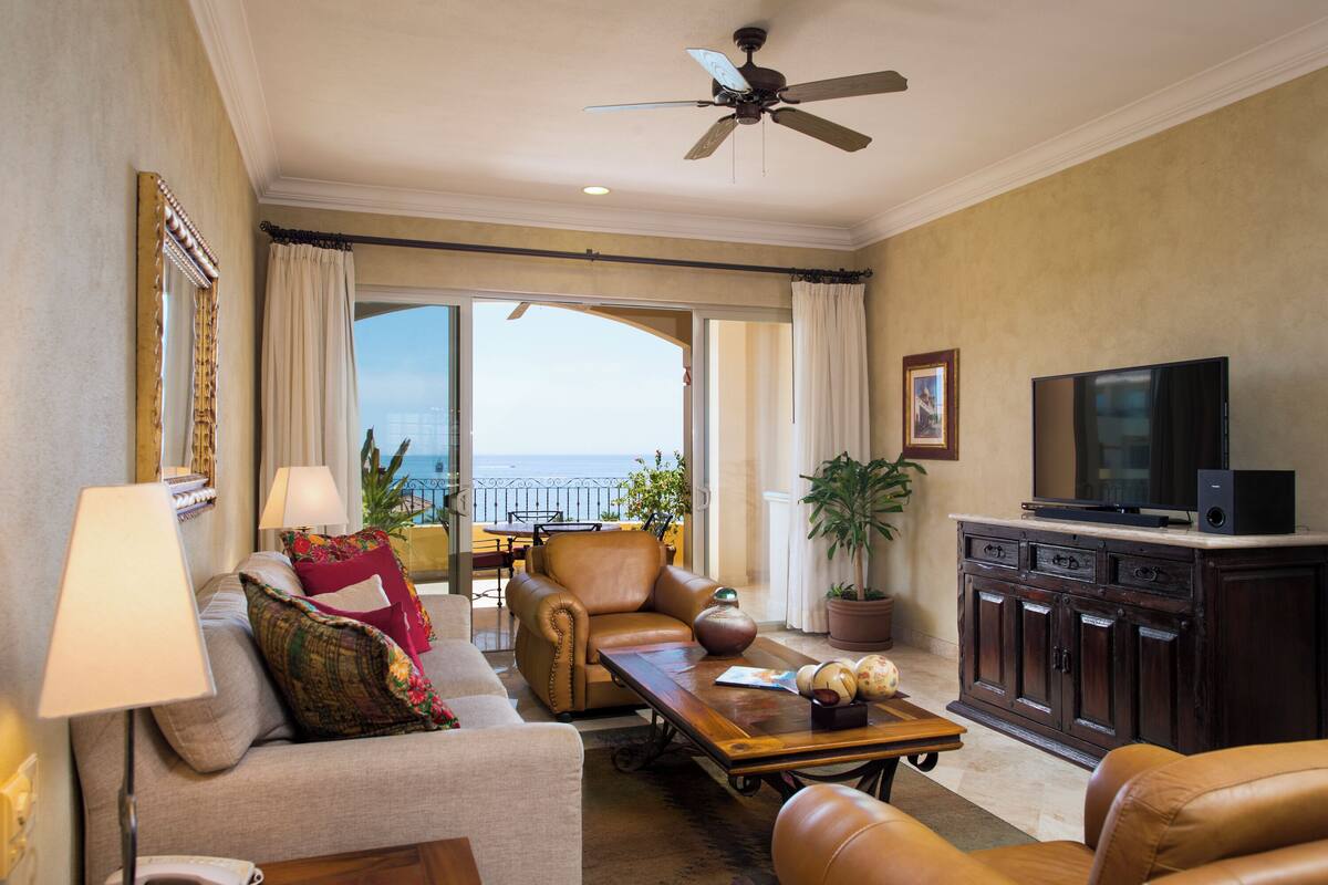 A spacious living room with a sofa, armchairs, a coffee table, and a television. There is a balcony with a view of the ocean.