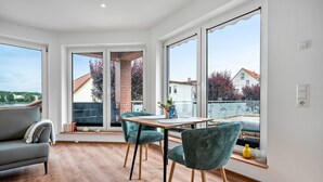 Una sala de estar moderna con un sofá, una mesa de comedor con sillas, ventanas amplias y vistas a edificios y árboles.