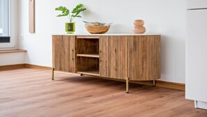 Un mueble de madera con un cuenco y un jarrón encima, una planta a su lado y un refrigerador blanco al fondo.