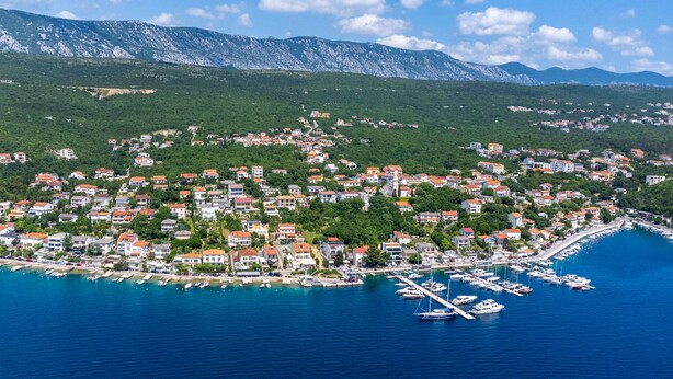 A coastal town with a marina, houses with red roofs, and mountains in the background.