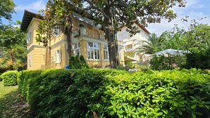 A yellow building with balconies, surrounded by lush greenery and a clear sky. - Apartman Villa Adelle - Lovran by Villas Guide (Lovran)