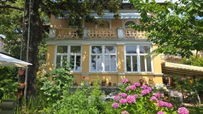 Een geel gebouw met veel ramen en balkons, omgeven door weelderige begroeiing en roze bloemen.
