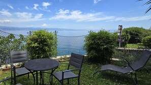 A patio with a table and chairs overlooking a body of water, surrounded by greenery and a chain-link fence.