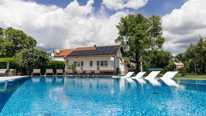 En swimmingpool med solstolar, ett hus med solpaneler på taket och träd i bakgrunden.