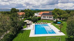 A swimming pool with a wavy edge, surrounded by a well-maintained lawn, a patio with outdoor furniture, and a house with solar panels.