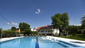 Ein Schwimmbad mit Liegestühlen, ein Haus mit rotem Ziegeldach und umgebende Grünflächen.
