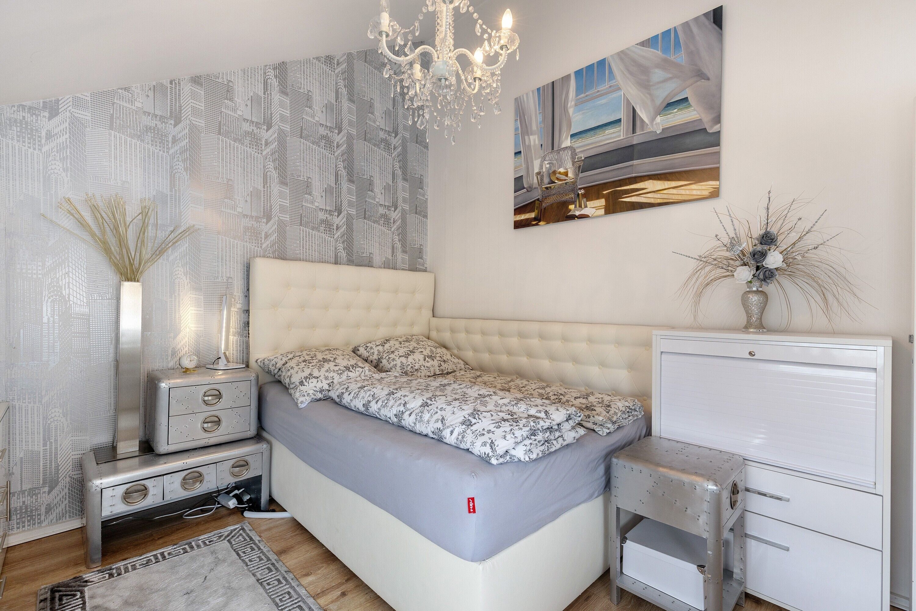 A bedroom with a bed, nightstands, a dresser, a chandelier, and a cityscape wallpaper.