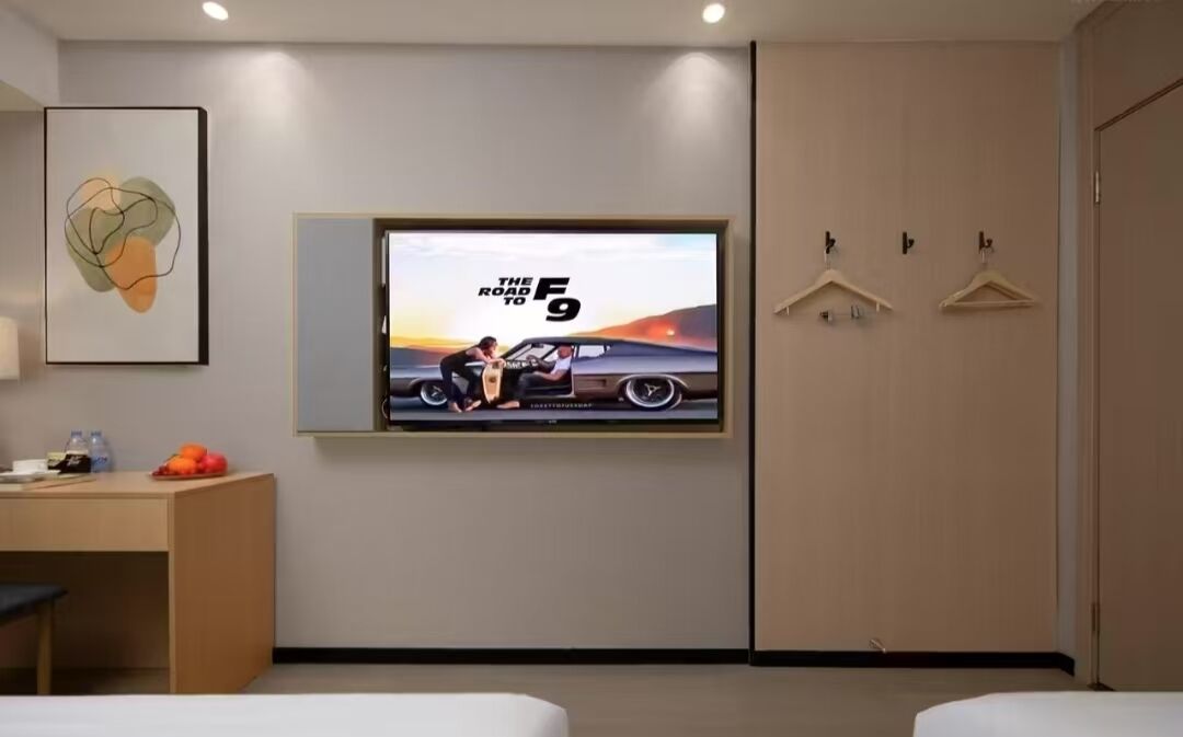 Habitación de hotel con una televisión de pantalla grande mostrando un cartel de película, un escritorio con una silla, una cama y un armario con perchas.