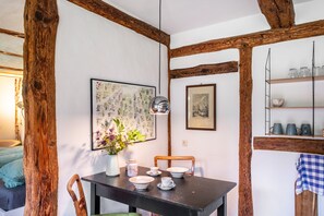 Un coin repas rustique avec des poutres apparentes, une table dressée avec des assiettes et des tasses, un vase rempli de fleurs et, au mur, des reproductions encadrées de gravures botaniques.