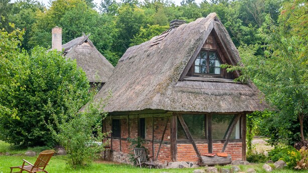A rustic thatched-roof cottage with a brick chimney, surrounded by lush greenery. - Holiday Home 'Im Grünen' in the Leonorenwald, Near the Baltic Sea (Borkenhagen / Kalkhorst)