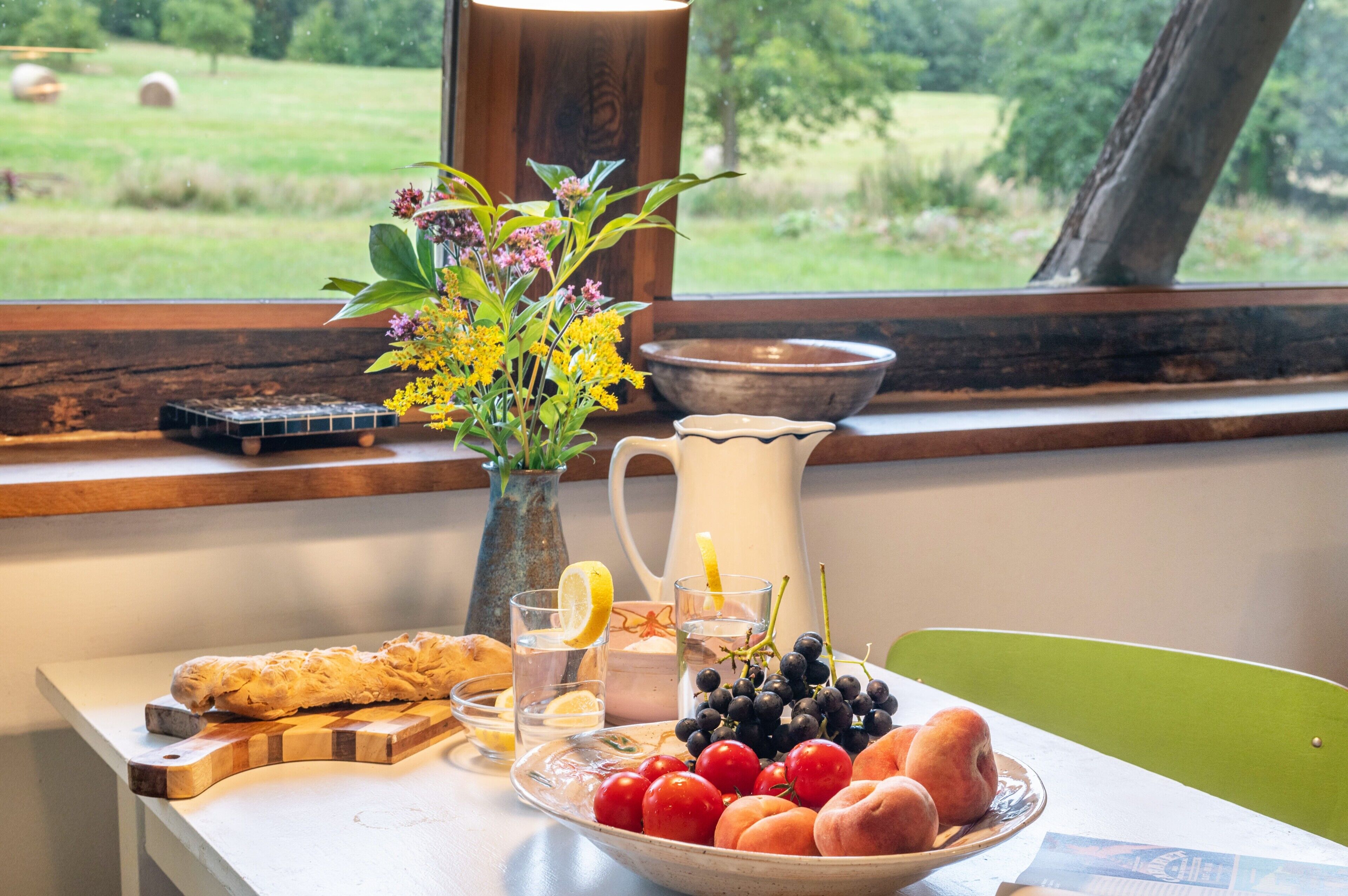 A breakfast table with bread, fruit, a vase with flowers, a pitcher, and a view of a green landscape.