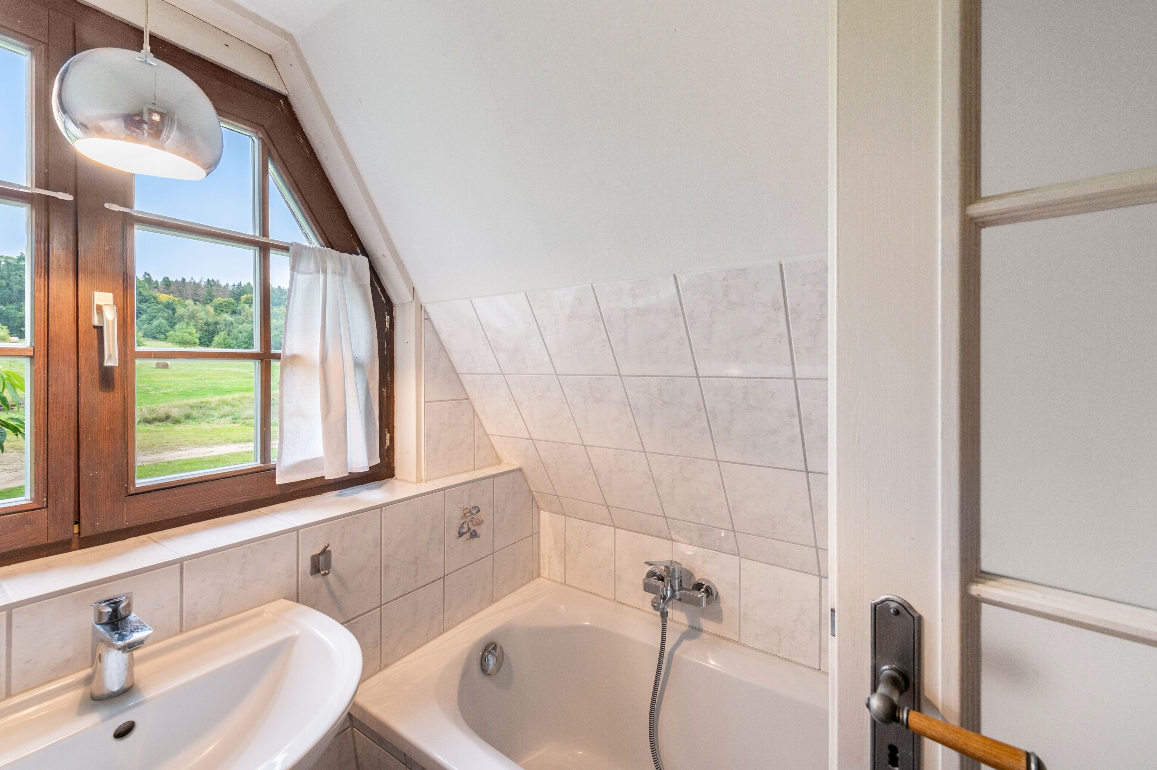 Ein Badezimmer mit einer Badewanne, einem Waschbecken, einem Fenster mit Vorhängen und Blick ins Grüne.