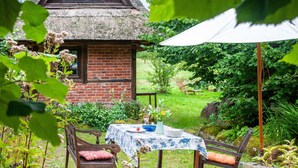 A rustic wooden table with a white tablecloth, surrounded by a thatched-roof building, a bench, and a garden setting. - Holiday Home 'Im Grünen' in the Leonorenwald, Near the Baltic Sea (Borkenhagen / Kalkhorst)