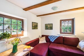 Un cuarto acogedor con un sofá rojo, una mesa de madera con un jarrón de flores y dos ventanas con vistas al verdor.