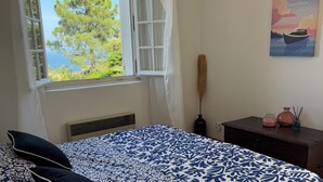 A bedroom with a bed, a window with a view of trees and the sea, a nightstand with a vase, and a wall with a poster.