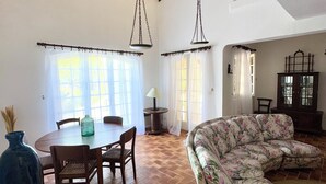 A spacious living area with a large floral sofa, a dining table with chairs, and a wooden floor.