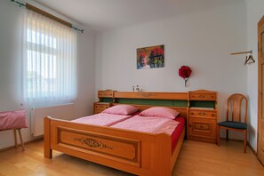 A bedroom with a wooden bed, a chair, a radiator, a window with sheer curtains, a picture on the wall, and a door. - Apartment 'Renchglück' with Shared Terrace, Shared Garden and Wi-Fi (Renchen)