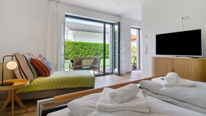 A hotel room with a bed, a TV, a seating area with a bench, and a view of a green hedge and building outside.