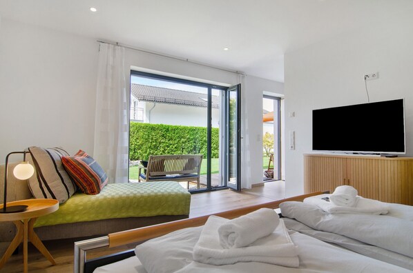 A hotel room with a bed, a TV, a seating area with a bench, and a view of a green hedge and building outside.