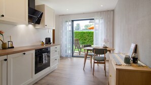 A modern kitchen with a dining area, a TV, and a view of a green hedge.