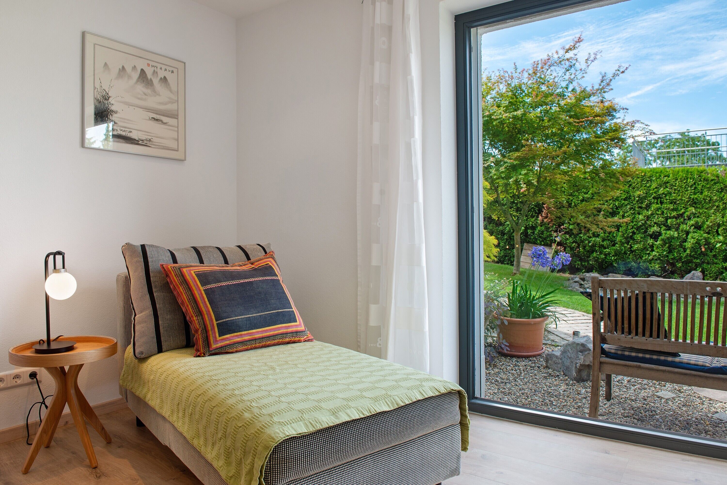 Een slaapkamer met een bed, nachtkastje, lamp en uitzicht op de tuin door een raam.