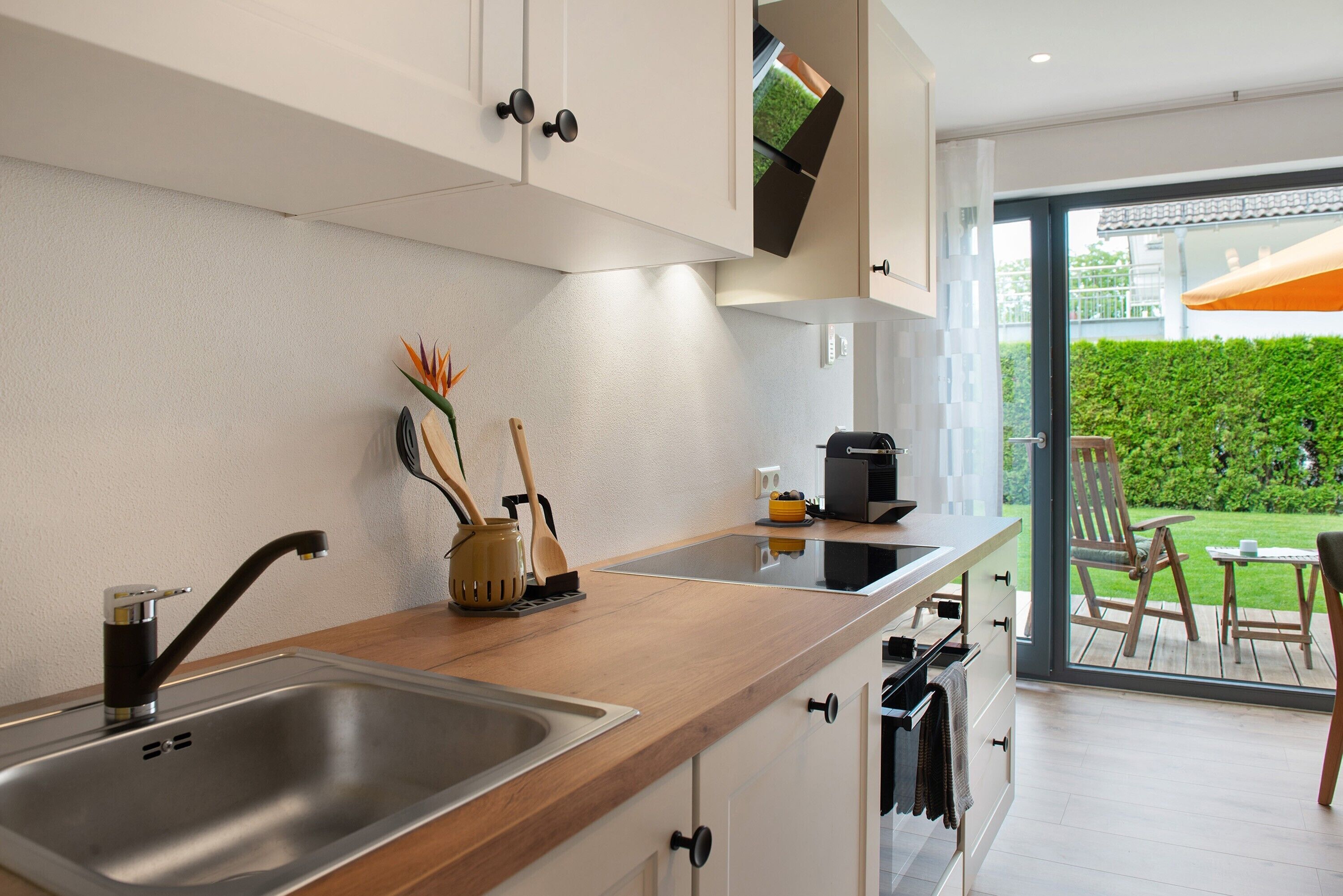 Une cuisine moderne dotée d’un évier en acier inoxydable, d’un plan de travail en bois et offrant une vue sur un jardin verdoyant grâce à une porte vitrée.