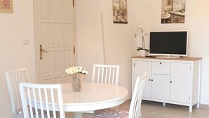 A dining area with a round table, four white chairs, a chandelier, and a cabinet with a television.