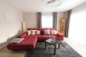 Un salon moderne avec un canapé d’angle rouge, une petite table avec des bougies, un élément décoratif mural et une grande horloge en bois.