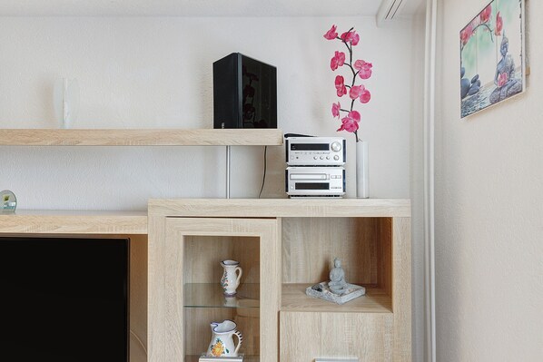 Una habitación con un escritorio de madera, un televisor negro, un altavoz blanco, un jarrón decorativo y una estatua de Buda.