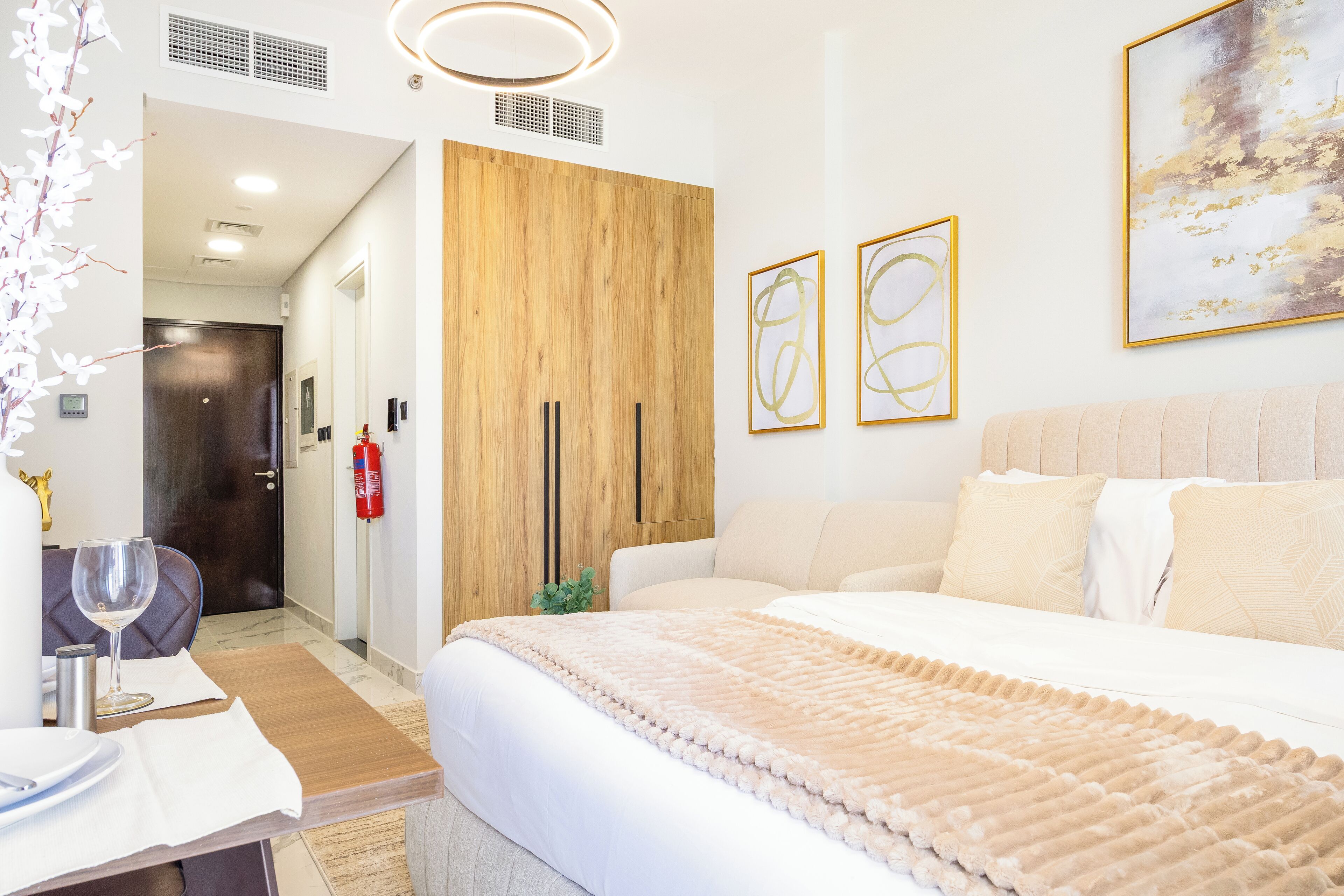 A hotel room with a wooden wardrobe, a bed with a beige blanket, a dining table with a wine glass, and two framed abstract paintings on the wall.