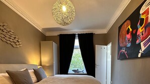 Une chambre avec un grand lit, un luminaire suspendu, une fenêtre avec des rideaux et un tableau au mur.