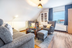 A modern living room with a sofa, armchair, ottoman, and a glass-top coffee table. There is a cabinet with glass doors, a lamp, and a view through a window. - Apartment 'Apfelkorb' with Private Terrace and Wi-Fi (Jork)
