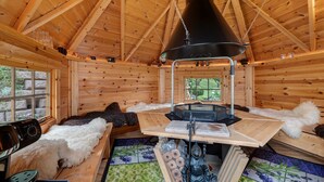 L’intérieur d’une cabane en rondins, avec une table centrale, des murs en bois, une grande lampe noire et une fenêtre donnant sur un paysage.