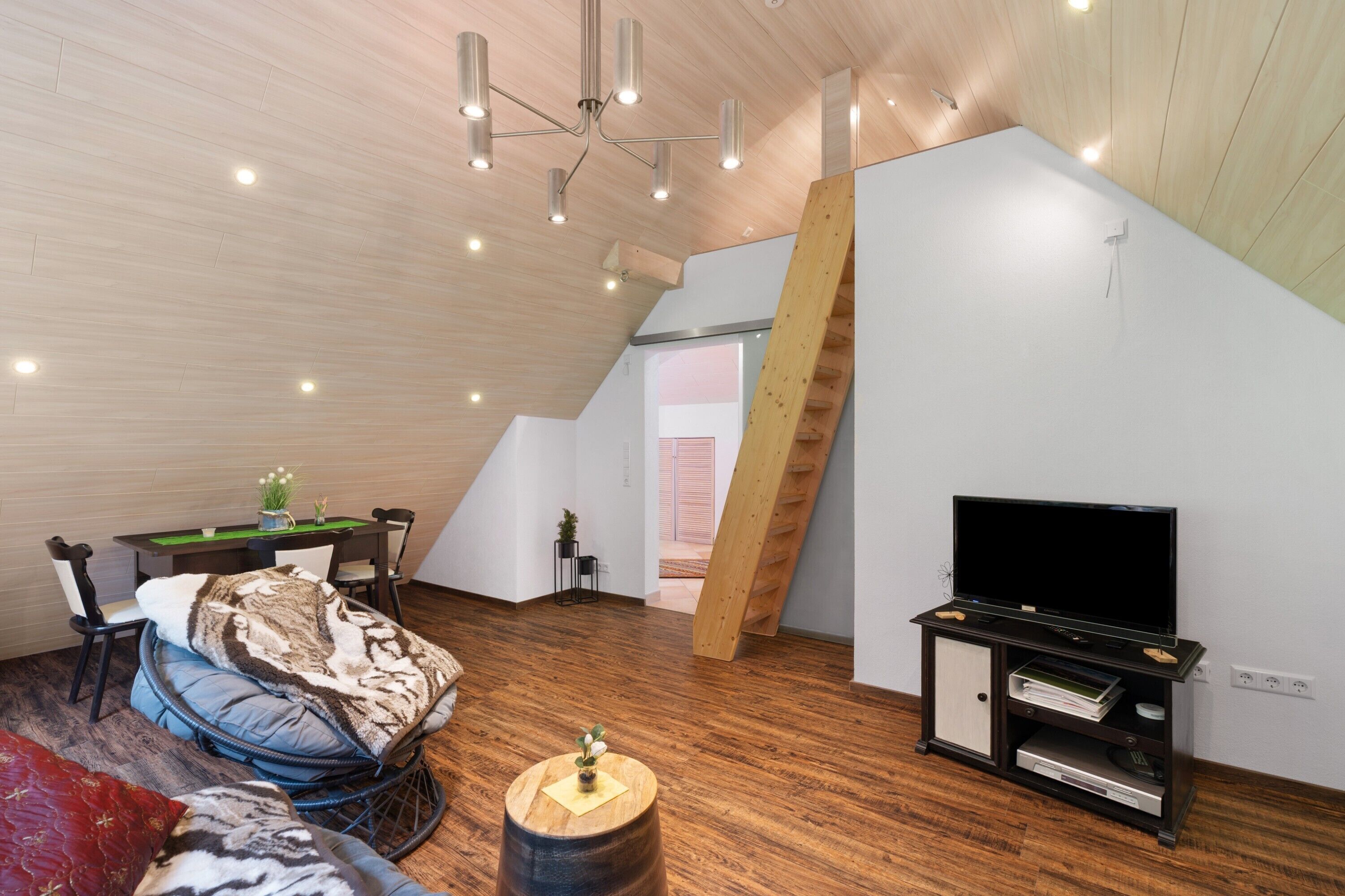 A loft-style room with a wooden floor, a TV on a black stand, a dining table with chairs, a sofa, and a staircase.