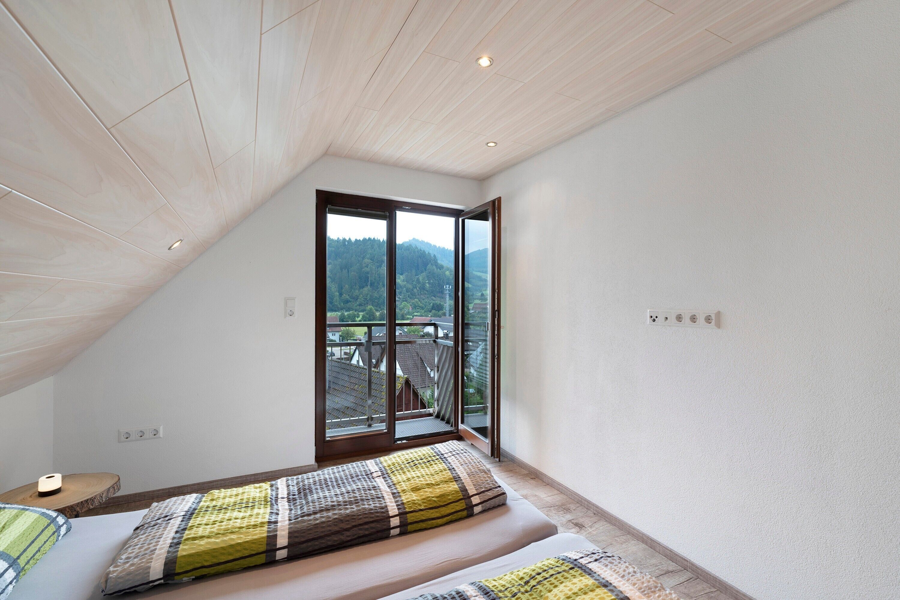 Ein Schlafzimmer mit schrägen Wänden, einem Bett mit gemustertem Überwurf, einem Nachttisch und einem Fenster mit Blick auf eine bewaldete Landschaft.