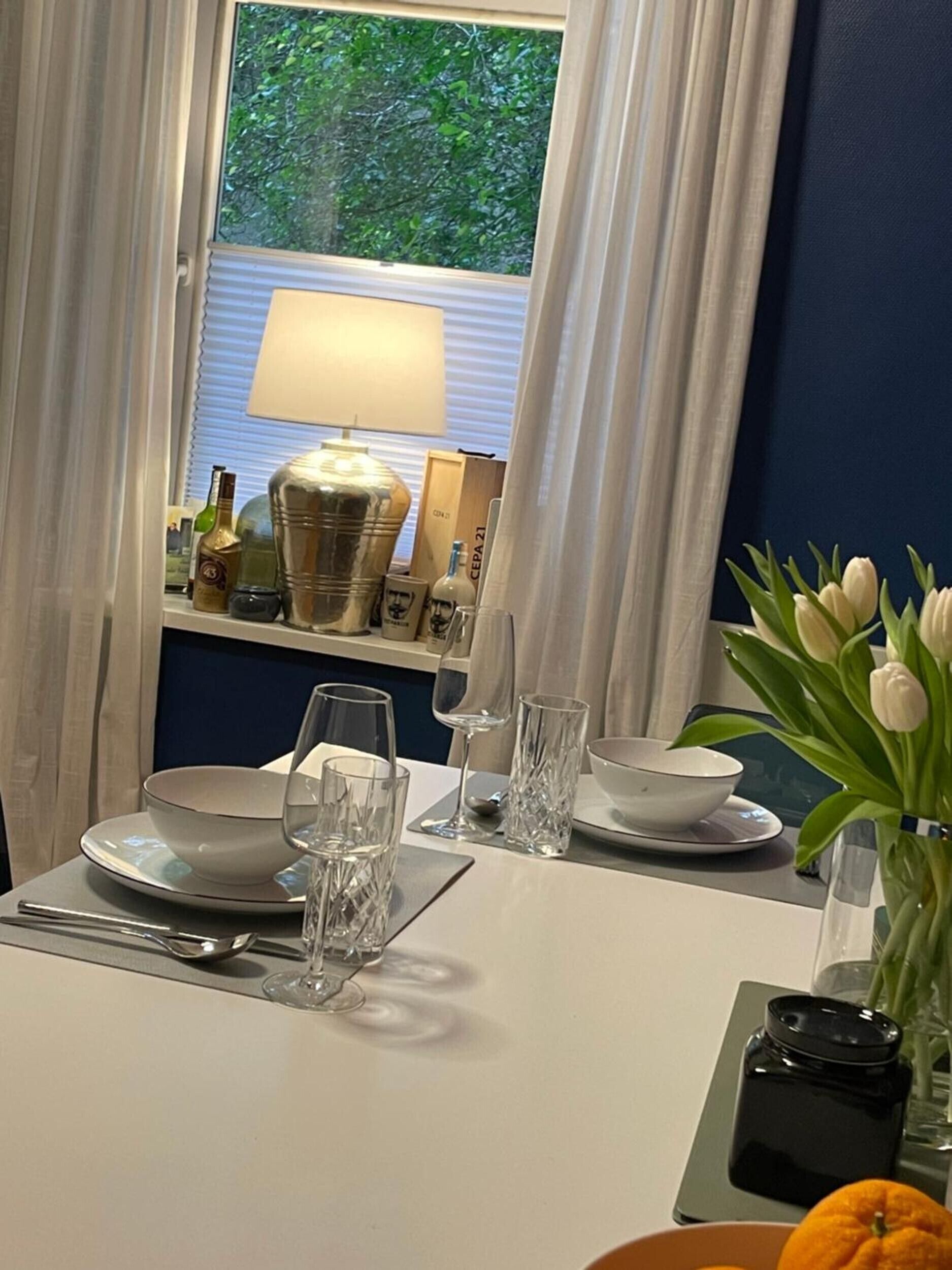 A dining table set with plates, glasses, and cutlery, a vase with flowers, and a window with curtains.