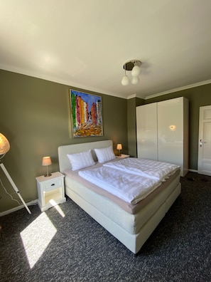 Ein ordentlich bezogenes Bett mit weißen Bettwäsche, ein Gemälde von Gebäuden an der Wand, ein Kleiderschrank und eine Lampe auf einem Nachttisch.