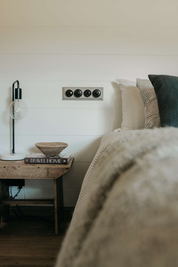 A cozy bedroom with a bed, a bedside table with a lamp, a book titled "TRAVEL HOME," and a wall-mounted air vent.