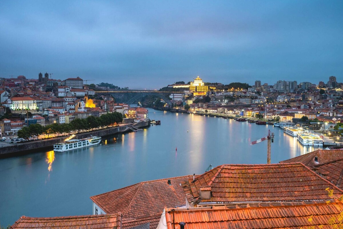 Paesaggio urbano lungo un fiume, con edifici storici, un edificio giallo ben illuminato e un ponte.