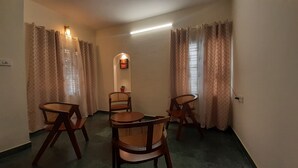 A room with a round table and four wooden chairs, beige curtains, a small potted plant, and a wall-mounted light fixture.