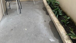 Un petit espace extérieur comprenant une table et une chaise en métal, un sol en béton et un bac à fleurs surélevé où sont plantées des plantes vertes.