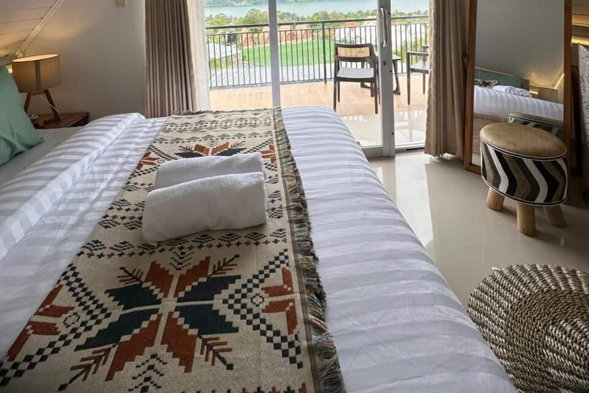 A bedroom with a large bed, a mirror, a view of the outdoors, and a balcony.