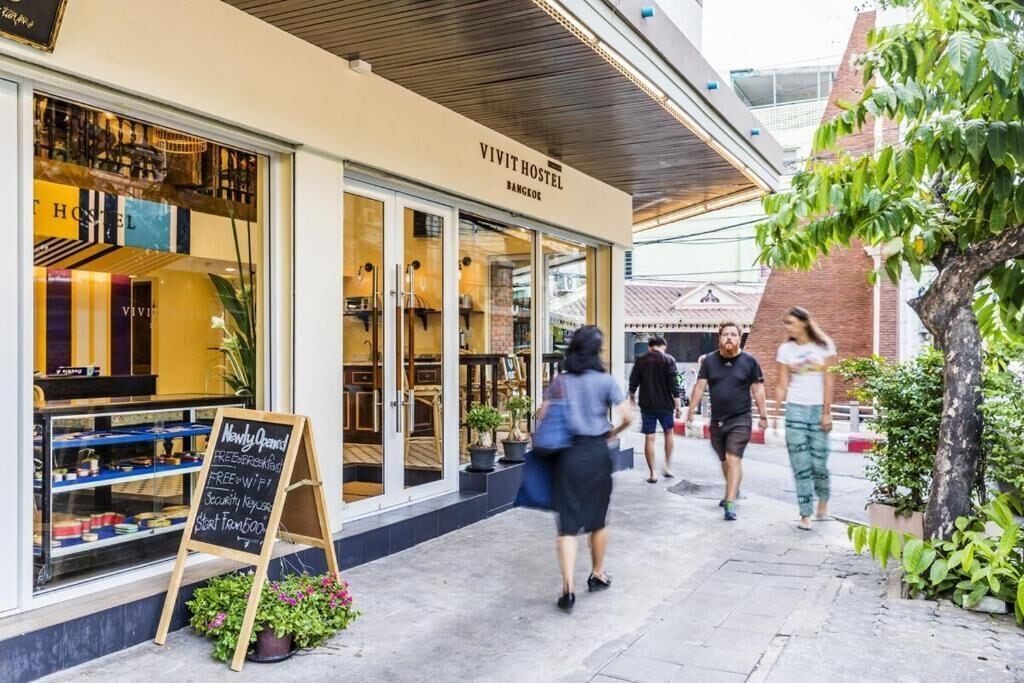 Un hostal Vivit en Bangkok con fachada moderna, ventanas grandes y un cartel anunciando nuevas inauguraciones y desayuno.